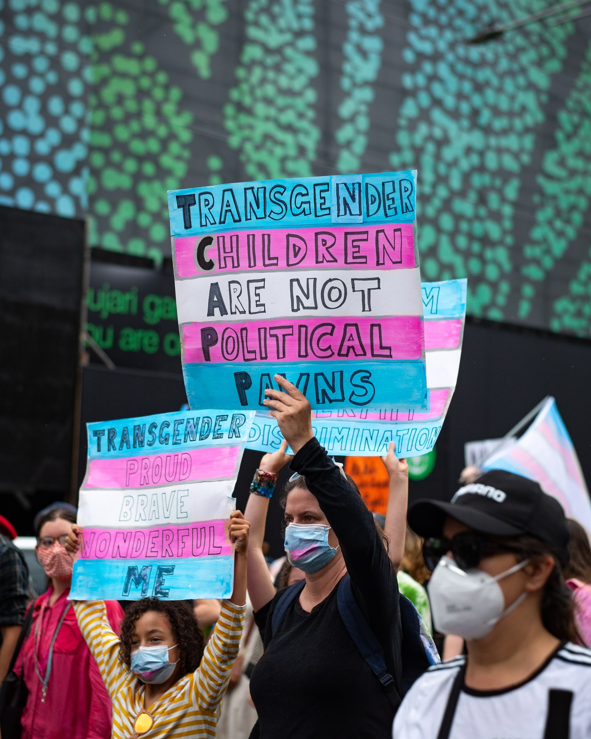A group of people holding signs that read, Transgender Children are not Political Pawns, and wearing masks photo.
