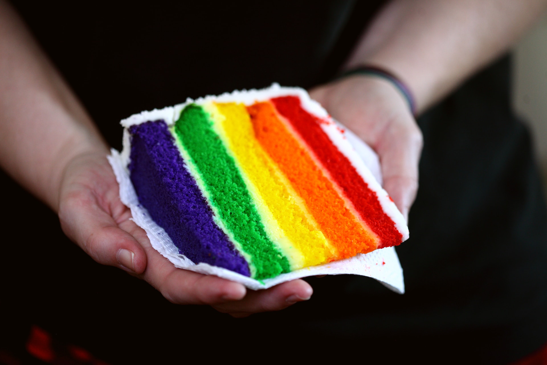 Two hands holding a slice of rainbow cake.