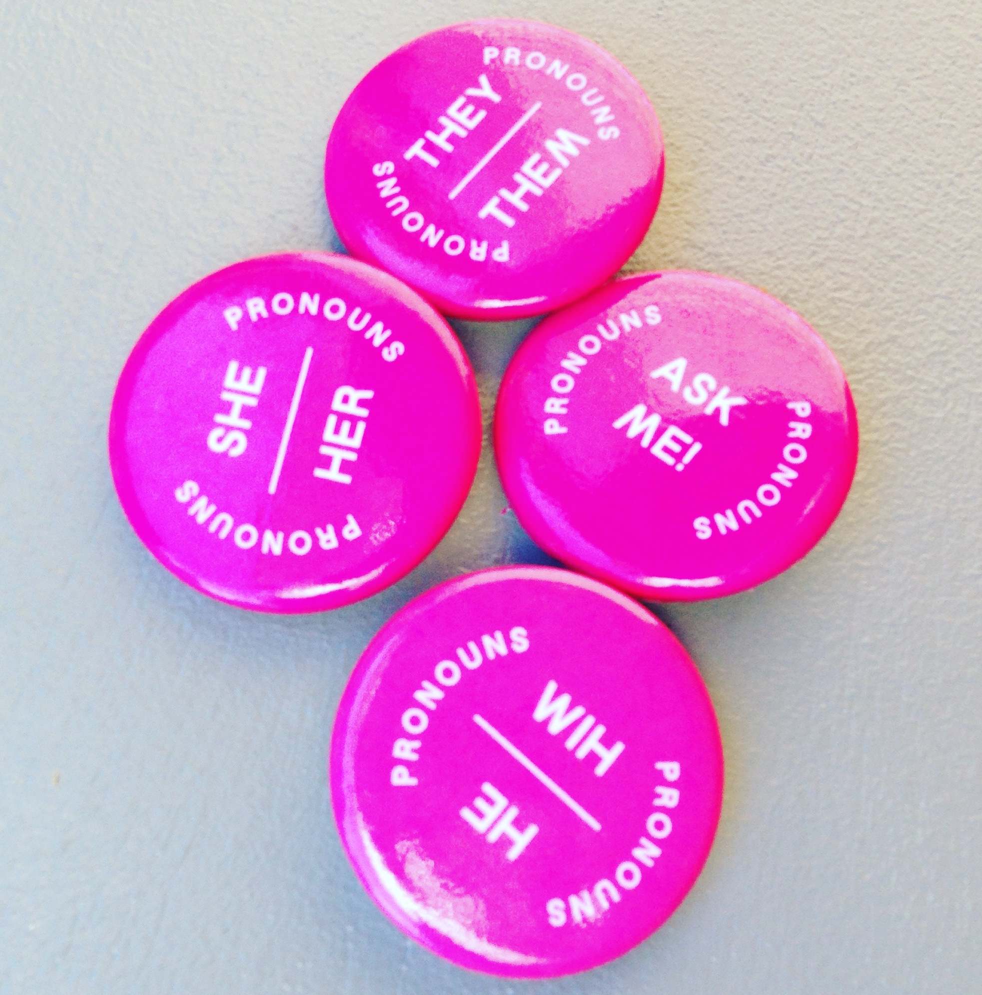 Four pronoun pins with the various pronouns on them and one that states Ask Me! about my pronouns.