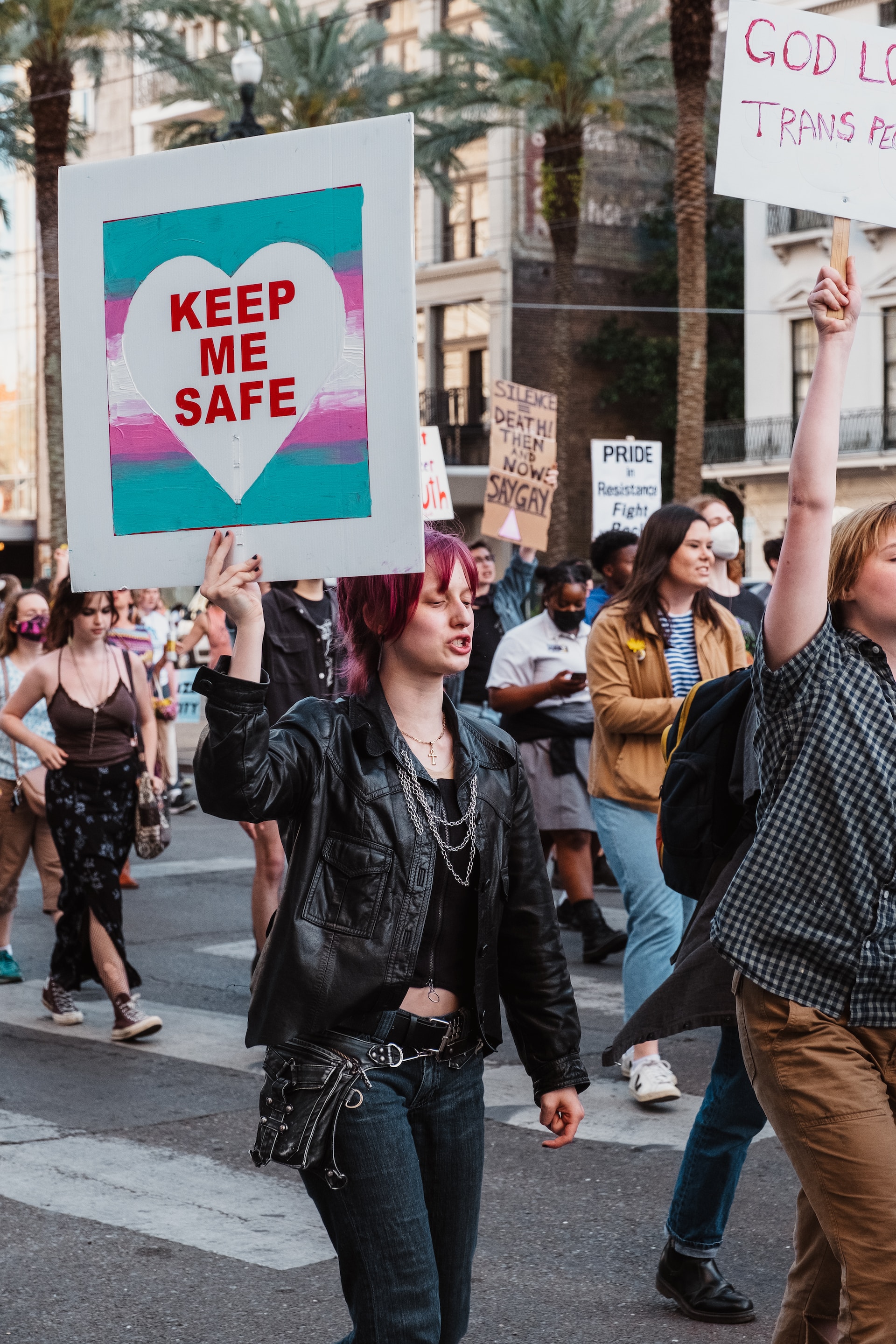 A photo of a crowd of people marching down the street holding signs: one reads, Keep me Safe, and another reads, God loves Trans people.